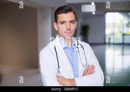 Médecin homme croisant les bras dans le couloir de l'hôpital portant une chemise bleue, une blouse de laboratoire et un stéthoscope Banque D'Images