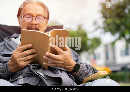 Homme asiatique âgé avec des lunettes lisant un livre à l'extérieur en automne près d'un arbre, regardant réfléchi. Copier l'espace. Concept de lecture senior. Banque D'Images