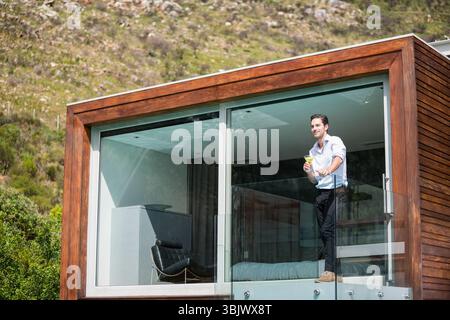 homme debout sur un balcon à balustrade de verre tenant un verre surplombant une colline herbeuse, à côté d'une maison en bois Banque D'Images