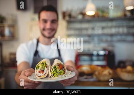 Barista portant un tablier tenant deux tortilla roulés avec laitue et tomate dans l'assiette au comptoir du café Banque D'Images