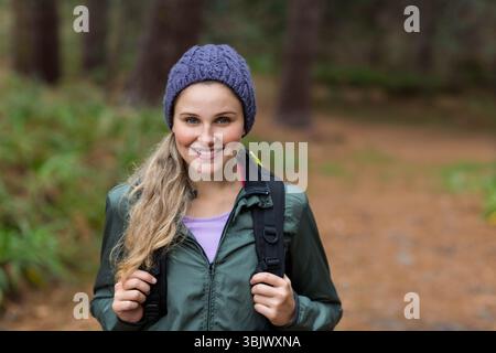 Femme debout sur le sentier forestier portant un bonnet violet et une veste verte tout en tenant des sangles de sac à dos Banque D'Images