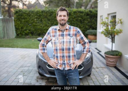 Homme dans la trentaine debout avec confiance sur l'allée de banlieue avec voiture compacte grise Banque D'Images