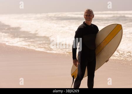Homme senior debout jusqu'au genou à la plage tenant une planche de surf et portant une combinaison, espace copie Banque D'Images