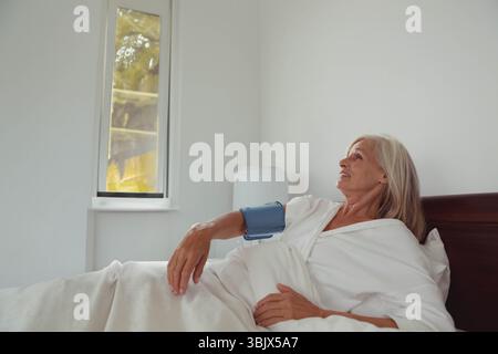 Femme âgée allongée sur le lit dans la chambre à coucher portant une robe d'hôpital et surveillant le brassard de tension artérielle Banque D'Images