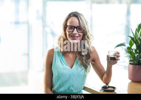Femme assise au comptoir dans le café sirotant du café de tasse en verre clair à côté des biscuits et de la plante Banque D'Images