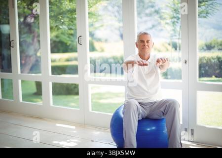 Homme senior équilibrant sur la balle de stabilité bleue dans la salle de gym à domicile par des portes en verre à cadre blanc, espace de copie Banque D'Images
