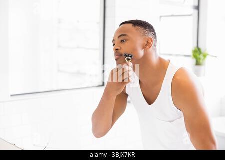 Homme afro-américain rasant le visage à l'évier de salle de bain devant le miroir avec rasoir de sécurité à main Banque D'Images