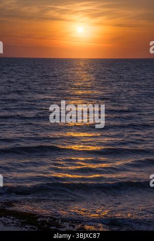 Le coucher de soleil vibrant peint le ciel avec des teintes chaudes tandis que de douces vagues ondulent dans l'océan, créant une atmosphère sereine. Banque D'Images