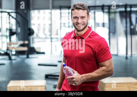 Homme musclé portant un polo rouge et un pantalon sombre tenant une bouteille d'eau à l'intérieur de la salle de gym près des boîtes plyométriques Banque D'Images