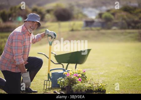 Homme senior agenouillé sur la pelouse arrière portant chapeau seau, utilisant la fourche à côté de brouette, espace de copie Banque D'Images