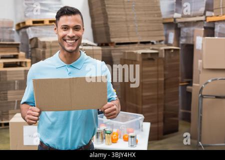 Ouvrier d'entrepôt masculin tenant le signe de carton vierge dans l'entrepôt au milieu des boîtes aplaties et du conteneur Banque D'Images