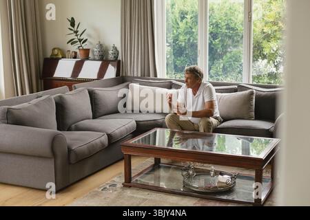 Homme senior assis sur le canapé dans le salon faisant des gestes vers la table basse en verre, espace de copie Banque D'Images