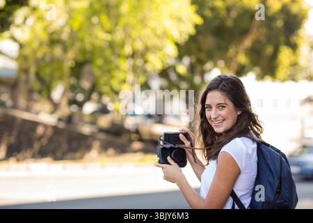 Femme debout à côté de la rue urbaine tenant un appareil photo rétro avec sac à dos bleu regardant en arrière, espace de copie Banque D'Images