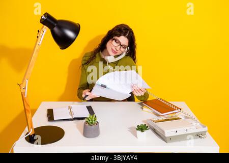 Femme occupée travaillant à un bureau avec des documents, un téléphone et une lampe, placée sur un fond jaune vif moderne Banque D'Images
