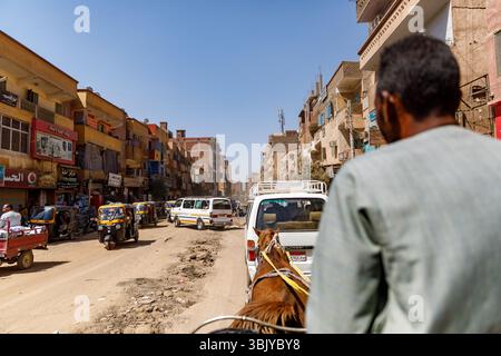 Une calèche tracée par des chevaux navigue dans une rue animée d'une ville égyptienne, partageant la route avec des pousse-pousse et des fourgonnettes automobiles, mettant en valeur un mélange de transport traditionnel et moderne. Banque D'Images