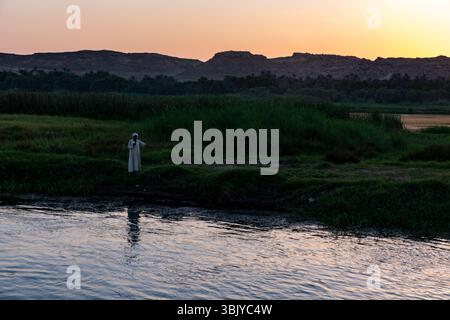 Un homme vêtu de vêtements traditionnels se tient au bord de la rivière, peut-être pêchant ou observant le paysage au crépuscule. Banque D'Images