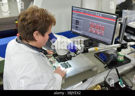 Femme dans une usine pour la production de composants électroniques vérifie la qualité d'un conseil assemblé avec l'aide d'un microscope ou une loupe gl Banque D'Images
