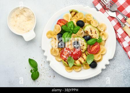 Salade de pâtes fiocchi avec légumes courgettes grillées, tomates cerises, olives, basilic et fromage parmesan dans une assiette blanche sur lumière s. Banque D'Images