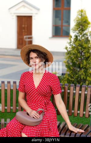 Femme souriante en robe enveloppée rouge et chapeau pose par bâtiment. Banque D'Images
