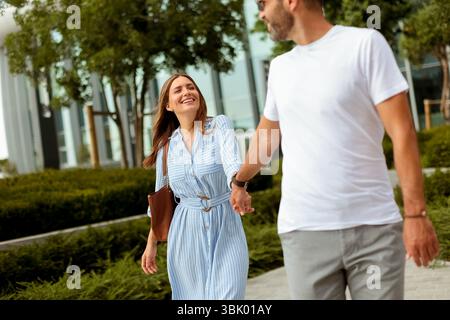 Un couple se promène main dans la main avec des sourires sur le visage, entouré de verdure luxuriante dans un parc urbain animé par une journée lumineuse. Banque D'Images