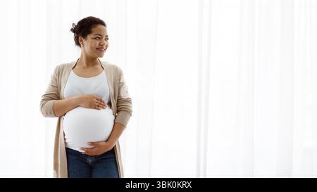 Belle femme enceinte noire debout près de la fenêtre à la maison, embrassant le ventre Banque D'Images