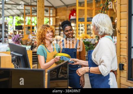 Client payant avec paiement sans contact dans une épicerie, assisté par deux vendeurs souriants Banque D'Images