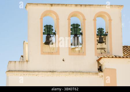 Trois cloches en bronze vieillies sont accrochées dans les arches aux finitions pastel d'une tour d'église historique contre un ciel bleu clair à Tavira, au Portugal. Banque D'Images