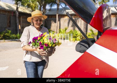 Femme âgée heureuse portant des pétunias colorés dans un pot au coffre ouvert de sa voiture rouge, prête pour une excursion d'une journée ensoleillée Banque D'Images