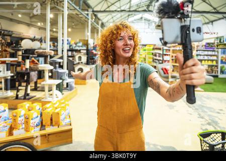 Jeune vendeuse filmant une vidéo pour les médias sociaux avec un smartphone et un microphone, montrant des produits pour animaux de compagnie dans une animalerie Banque D'Images