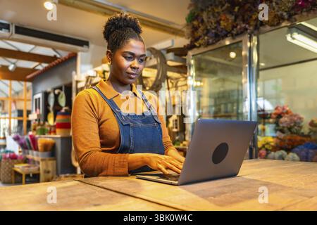 Jeune fleuriste gérant les commandes en ligne à l'aide d'un ordinateur portable dans son magasin de fleurs, entouré de fleurs et de plantes colorées Banque D'Images