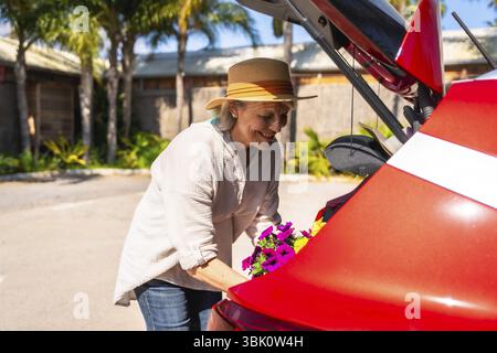 Heureuse femme âgée plaçant soigneusement des pétunias colorés dans le coffre de sa voiture rouge, prête à les ramener à la maison Banque D'Images