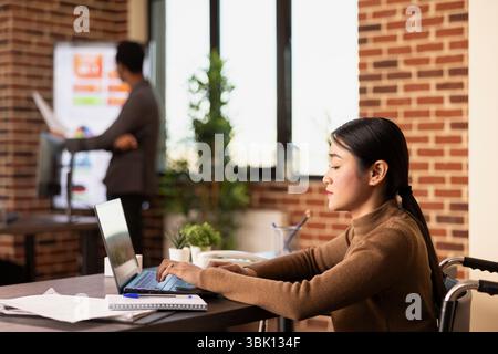Employé féminin asiatique dans le dactylographie en fauteuil roulant sur ordinateur portable, la recherche d'analyses d'affaires dans le bureau de mur de briques. Gestionnaire de femme focalisée utilisant un appareil numérique au bureau, travaillant sur le développement d'un projet d'entreprise. Banque D'Images