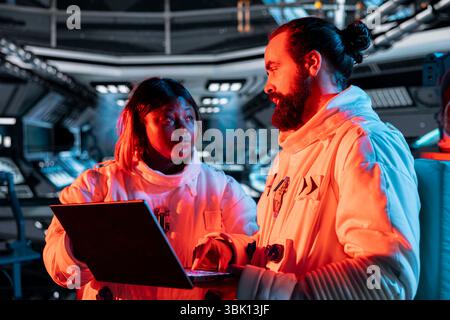 Les ingénieurs spatiaux travaillant en équipe utilisent un ordinateur portable pour réparer une brèche de sécurité provoquant un avertissement de surcharge. Collègues de la station spatiale utilisant un ordinateur portable, réparant une erreur système critique déclenchant des lumières de secours Banque D'Images