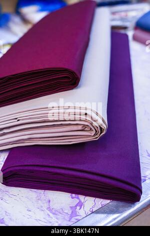 Trois rouleaux de tissu soigneusement pliés dans les tons de violet, Bordeaux et beige sont affichés sur une table. Les rouleaux sont empilés, mettant en valeur le tex riche Banque D'Images