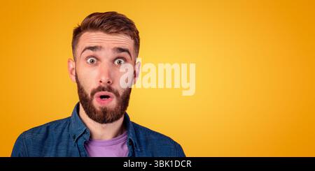Des nouvelles incroyables. Portrait de jeune barbu ouvrant la bouche et les yeux en choc, regardant l'appareil photo sur fond orange Banque D'Images
