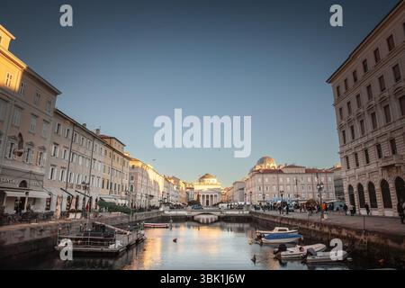 TRIESTE, ITALIE - 17 DÉCEMBRE 2024 : la lumière dorée du crépuscule se reflète sur les bateaux qui bordent Trieste canal Grande, avec l'église de Sant Antonio Taumaturgo. Cana Banque D'Images