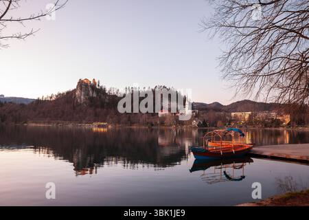 Vue au crépuscule sur le lac de Bled en Slovénie avec un château de Bled incandescent sur sa falaise et la flèche de l'église Saint-Martin reflétée dans l'eau calme, tandis qu'un pletna vide Banque D'Images