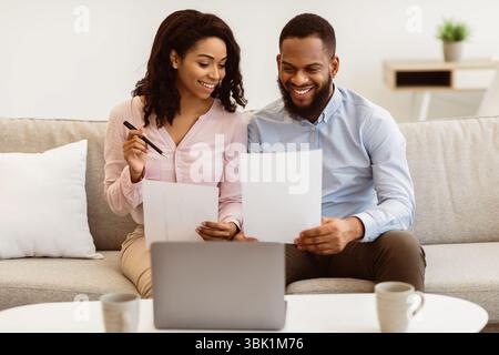 Couple noir joyeux lisant des documents à la maison, vérifiant des factures Banque D'Images