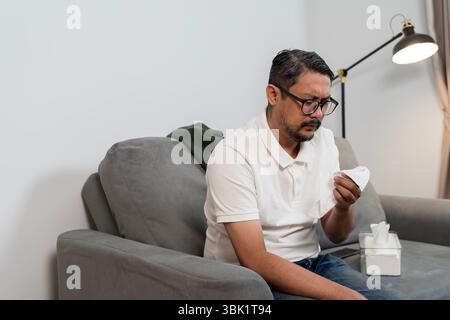 Un homme d'âge moyen portant des lunettes et une chemise blanche assis sur un canapé gris tenant un mouchoir avec une expression triste, peut-être se sentir malade ou émotionnellement vers le haut Banque D'Images