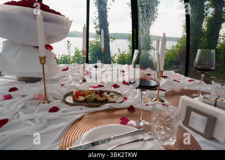 Salle à manger intime avec un bouquet de roses rouges, des verres à vin, des bougies allumées et un plateau de fromages sur une table couverte de blanc dans un overlooki de maison en verre Banque D'Images
