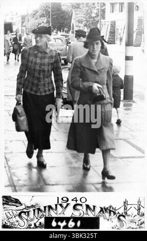 Londres, Angleterre. 1940 : photographie de rue de la seconde Guerre mondiale prise par Sunny Snaps en 1940. Il représente une femme d'âge moyen et une femme âgée marchant le long d'une rue londonienne non identifiée. L'un porte un sac à provisions et l'autre, un panier. De l'autre côté de la route se trouve un bâtiment avec des sacs de sable empilés devant lui pour se protéger contre les explosions de bombes. Les montants de lampe sont peints avec des bandes blanches pour faciliter la visibilité en cas de panne sèche. Banque D'Images