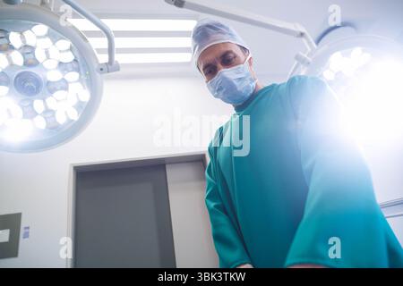 Chirurgien masculin debout dans les gommages sous les lampes dans la salle d'opération, inspectant le champ chirurgical Banque D'Images