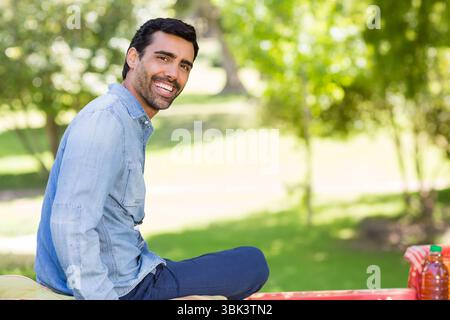 Homme adulte moyen assis sur le banc de pique-nique rouge souriant dans le parc avec des bouteilles de jus, espace de copie Banque D'Images