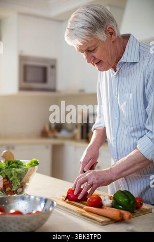 Homme senior debout dans la cuisine à la maison tranchant des légumes avec couteau de chef sur la planche à découper, espace de copie Banque D'Images