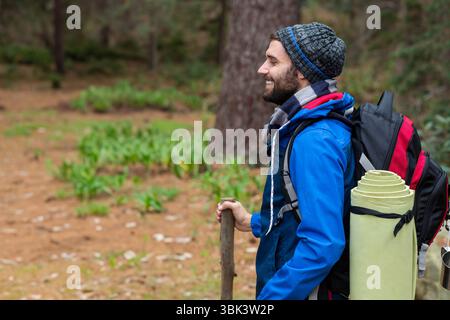 Homme randonnée dans la forêt de pins avec sac à dos, tenant un bâton de marche et portant un bonnet en tricot, espace de copie Banque D'Images