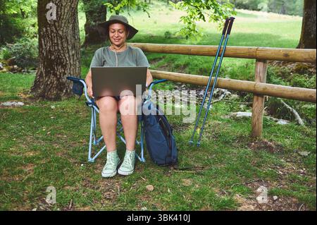 Une femme est assise dans un espace extérieur serein, travaillant sur son ordinateur portable. Son environnement naturel et son équipement de randonnée suggèrent son plaisir de la nature et une vie en plein air Banque D'Images