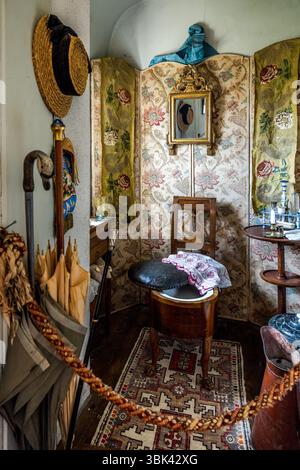 Une pièce avec un coin toilettes au Château de Boussac, département de la Creuse, Nouvelle-Aquitaine, France, est meublée d'une chaise de commodité (chaise percée) (peut-être la pièce dans laquelle George Sand a séjourné lors de ses visites). La chaise en bois se trouve devant un paravent recouvert d'un tissu à fleurs. Elle possède un dossier en forme de lyre, un coussin de siège noir et un bassin en porcelaine, avec un tissu à motif rouge drapé par-dessus. Un miroir doré est accroché au paravent. Un chapeau de paille, une canne à pommeau en forme de tête de canard et des parapluies pendent à gauche, et une table d'appoint ronde avec des bouteilles en verre et une bougie se trouve sur le Banque D'Images