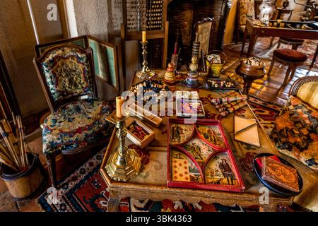 Le salon du Château de Boussac, département de la Creuse, Nouvelle-Aquitaine, France, présente une collection de jeux de société. Un plateau rouge pour le jeu de cartes et de hasard français Nain Jaune (« Nain Jaune ») avec cinq compartiments et des motifs de cartes tels que le sept et le dix de carreaux repose sur une table avec une nappe brodée. À côté se trouvent des pièces de jeu dans un bol, des toupies, des cartes à jouer, des boîtes de jeu et une petite ardoise avec un crayon, et deux chandeliers en laiton tiennent des bougies. À gauche, une chaise avec une nappe brodée se tient à côté d’un seau en bois avec des bâtons, et un abaque devant la cheminée Banque D'Images