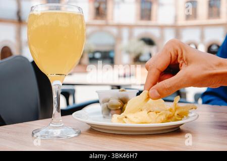 un homme est sur le point de manger une croustille de pomme de terre dans une assiette placée sur une table dans un café sur le trottoir quelque part en espagne, où il y a un verre avec un mélange de bière shandy Banque D'Images