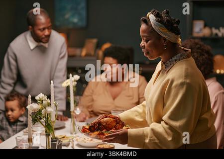 Femme noire servant de la dinde rôtie sur la table à manger avec des décorations florales tandis que la famille profite d'un dîner de vacances festif à la maison Banque D'Images